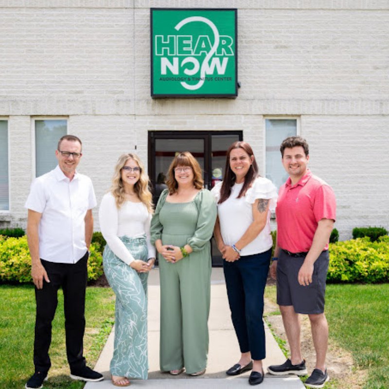 Angie Lederman with the Hear Now Audiology team outside the office, under the Hear Now sign